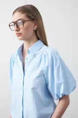 Camisa blanca de algodón con cuello camisero y mangas cortas abullonadas. Presenta un corte relajado y cierre frontal de botones.