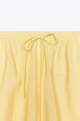 Pantalón de mujer color amarillo pálido, de corte ancho y tejido fluido. Tiene tiro medio, cintura elástica ajustable con cordones, bolsillos laterales y falsos bolsillos de vivo en la espalda.