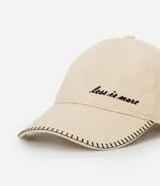 Gorra de algodón color beige con visera curva. Presenta bordado frontal en cursiva negra con la frase "less is more" y pespuntes contrastantes en negro a lo largo del borde de la visera. Cuenta con cierre ajustable en la parte posterior mediante hebilla metálica.
