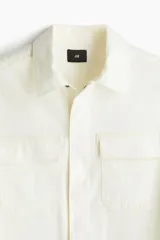 Sobrecamisa color crema con cuello inglés, cierre frontal con botones a presión, dos bolsillos superiores con solapa y puños ajustables con botón a presión.