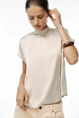 Blusa color crudo de tejido satinado, con cuello subido, manga sisa y cierre en la espalda con lazada y botones.
