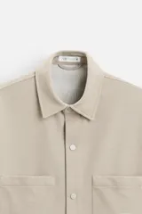 Sobrecamisa color beige de tejido elástico, con cuello solapa, manga corta y bolsillos de parche en el pecho. Cierre frontal con botones a presión.
