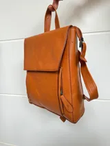 Mochila color marrón de cuero sintético, con asa de mano y correas ajustables. Tiene un bolsillo frontal con cierre a presión y un bolsillo lateral con cierre.