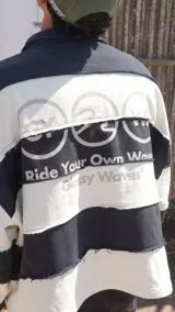 Buzo de tejido de punto grueso, estilo rugby, con cuello polo y tapeta de botones. Diseño a rayas horizontales anchas en blanco y azul marino, con bordes deshilachados en las uniones de las franjas. Presenta un logo bordado en el pecho.