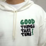 Canguro blanco con capucha, cordones ajustables y bolsillo tipo canguro. Estampado frontal con la frase "Good things take time" en verde y azul.