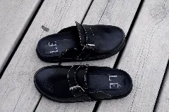 Zuecos negros de cuero con suela gruesa y correa ajustable con hebilla metálica plateada. Presentan costuras blancas visibles en la parte superior.