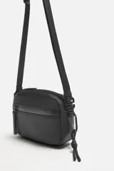 Bandolera negra de acabado engomado con correa ajustable y extraíble. Tiene un compartimento principal con cierre de cremallera, un bolsillo interior con cremallera y un bolsillo frontal con cremallera.