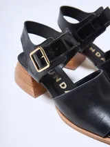 Sandalias de cuero negras con punta cuadrada, tira ancha sobre el empeine y pulsera con hebilla dorada al tobillo. Taco geométrico de madera clara.