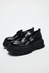 Zapato tipo mocasín negro con plataforma y suela track. Presenta tira ajustable adherente y tirador trasero.