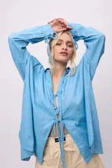 Camisa blanca de lino y algodón, con cuello clásico, cierre frontal con lazos y mangas largas abullonadas con puños ajustables con lazos.