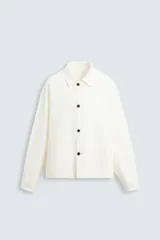 Sobrecamisa color crudo de corte recto, confeccionada en tejido denim de algodón con textura espiga. Tiene cuello solapa, manga larga con puños abotonados, bolsillos de vivo en la cadera y cierre frontal con botones marrones.