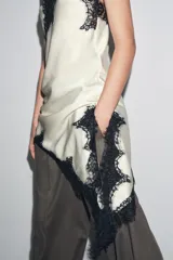 Vestido corto de satén color blanco, con tirantes finos, escote en pico y bajo asimétrico con abertura. Detalle de encaje negro en escote y ruedo.