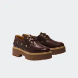 Zapato tipo náutico de cuero color marrón oscuro, con cordones y ojales metálicos dorados. Presenta una suela plataforma gruesa y dentada de color caramelo.