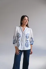 Camisa blanca de manga larga con elástico en puños, cuello mao con botones en la parte delantera y bordados florales azules en el frente y las mangas.