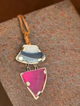 Collar con colgante de dos piedras naturales irregulares engarzadas en metal plateado y dorado, suspendidas de un cordón de cuero marrón con detalle de nudo corredizo. Las piedras tienen vetas en tonos naranja, marrón y blanco.