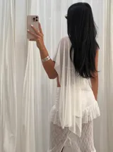 Capa asimétrica de tela negra transparente, tipo tul o gasa, con cuello halter y ojales metálicos. Se lleva sobre un top de bikini negro y una minifalda negra.