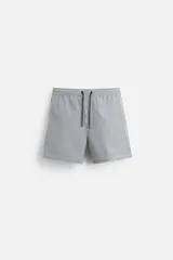 Short de baño con textura seersucker a rayas verticales azul marino y blanco. Tiene cintura elástica ajustable con cordón, bolsillos frontales y traseros, aberturas laterales en el bajo y forro interior.