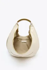 Bolso ovalado color crudo con asa de hombro metálica dorada. Tiene pespuntes en contraste, bolsillo interior con cremallera y cierre principal con cremallera y tirador.