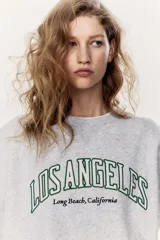 Buzo gris con cuello redondo, mangas largas y puños acanalados. Estampado frontal con el texto "Los Angeles Long Beach, California" en color verde.