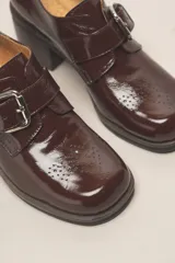 Zapato de cuero vacuno charol negro, con punta redondeada y detalle de perforado en la capellada. Tiene una tira con hebilla metálica y un taco cuadrado de 4 cm.