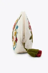 Neceser acolchado de forma redondeada, color crudo, con bordados florales grandes en tonos rojo y borgoña, y detalles de cinta verde. Posee un pequeño llavero/borla de hilo verde y rojo en el cierre.