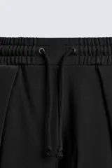 Pantalón jogger negro de corte relaxed fit, confeccionado en tejido de algodón. Presenta cintura elástica ajustable con cordón, pliegues frontales, bolsillos delanteros y un bolsillo trasero.