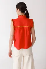 Blusa color mostaza sin mangas, con cuello alto y detalle de volados rojos en cuello, sisas y ruedo. Cierre frontal con botones.