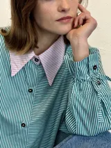 Camisa de popelina a rayas verticales verdes y blancas, con cuello rosa y botones marmolados. Es más corta adelante y más larga atrás.