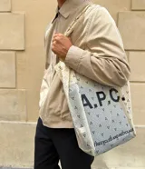 Bolso tote de tela color crudo con estampado de logos APC en negro y lunares azules. Tiene asas cortas y la inscripción "I am recycled and recyclable" en la parte inferior.