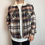 Chaqueta de franela a cuadros en tonos marrón, blanco, azul y naranja, con cuello de corderito blanco, cierre con botones a presión, bolsillos en el pecho con solapa y bolsillos laterales.