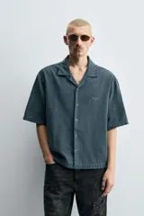 Camisa de denim de algodón, color beige claro, con corte relajado, cuello bowling y manga corta. Presenta cierre frontal con botones a presión y un pequeño bordado a tono en el pecho.