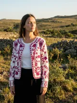 Chaqueta tipo cárdigan sin cuello, color blanco, con bordados florales intrincados en tonos magenta y fucsia. Presenta mangas largas y puños bordados.