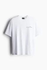 Remera blanca de algodón con cuello redondo y hombros caídos. Tiene un corte holgado y un pequeño estampado con la frase "Less is More" en el lado izquierdo del pecho.