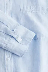 Camisa celeste de lino liviano, con cuello, botones al frente, canesú de doble capa en la espalda, hombros caídos, mangas largas con puños abotonados, bolsillo superior de parche y bajo levemente redondeado.