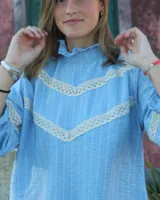 Camisa celeste con rayas verticales blancas y detalles de encaje color crema en forma de V en el frente y en los puños. Tiene cuello alto con volados.