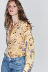 Camisa de ramio color beige con estampado floral en tonos marrones y azules, cuello solapa, manga larga con puños abotonados, bajo asimétrico con aberturas laterales y cierre frontal con botones.
