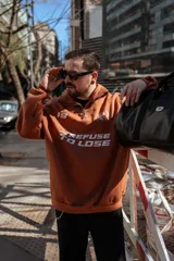 Buzo marrón con cierre medio, cuello alto y corte oversize. Estampado frontal con el número 19, un ancla con laureles y la frase "Refuse to Lose".