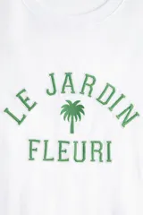 Remera blanca de algodón con corte amplio, cuello redondo y mangas cortas. Estampado frontal con texto en francés "Le Jardin Fleuri" y una palmera.