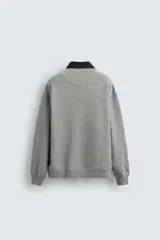 Sudadera gris de corte holgado, con cuello polo negro, cierre frontal de tres botones y mangas largas con puños acanalados.