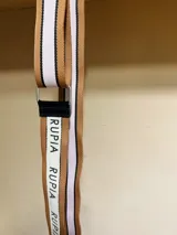 Correa para cartera de tela color marrón con rayas blancas y negras a los costados. Tiene la palabra "RUPIA" estampada en blanco sobre una franja blanca.