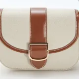 Bolso bandolera de lona color beige con detalles en cuero marrón. Tiene correa larga ajustable y cierre con hebilla dorada.