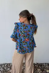 Blusa celeste con estampado floral en tonos naranja, verde y marrón. Tiene escote redondo y mangas cortas con volados.