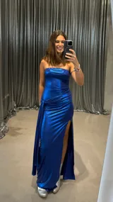 Vestido largo de fiesta azul eléctrico con textura brillante, corte ajustado, escote strapless y abertura lateral en la falda. Presenta un drapeado lateral con nudo.