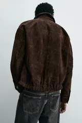 Cazadora de hombre de corte relajado confeccionada en piel con acabado ante, color marrón oscuro. Presenta cuello solapa, manga larga con puños abotonados a presión, bolsillos de vivo en cadera, bolsillo interior y bajo con elástico. Cierre frontal oculto por solapa con botones a presión.