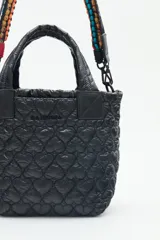 Cartera tote azul de nylon matelassé con diseño de corazones. Tiene manijas cortas y dos correas desmontables, una de cuero sintético y otra de tela trenzada multicolor. Cuenta con cierre superior y dos bolsillos internos.