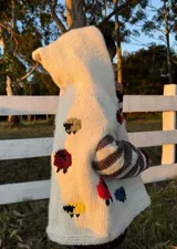 Poncho tejido a mano en lana merino color crudo, con capucha y mangas largas a rayas horizontales en tonos marrón y gris. Presenta apliques bordados de ovejas de colores.