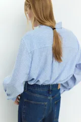 Camisa celeste y blanca a rayas verticales, confeccionada en mezcla de lino y viscosa. Tiene cuello clásico, botones frontales, bolsillo de parche en el pecho, mangas largas con puños abotonados y bajo redondeado.
