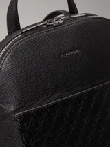 Mochila negra de piel sintética granulada tipo caviar, con bolsillo exterior con textura de logo monograma y logo de metal plateado brillante en la parte delantera. Tiene asa de mano en la parte superior, correa para el hombro ajustable, cierre de cremallera en la parte superior con logo brillante en los tiradores y bolsillos interiores con ranura.