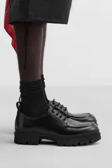 Zapato de vestir tipo blucher color negro, con detalle de bordón, tirador trasero y horma redonda. Suela con volumen y diseño irregular.