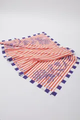 Pareo de algodón con estampado de rayas verticales naranjas y blancas, con diseño floral en azul y bloques violetas en los extremos.